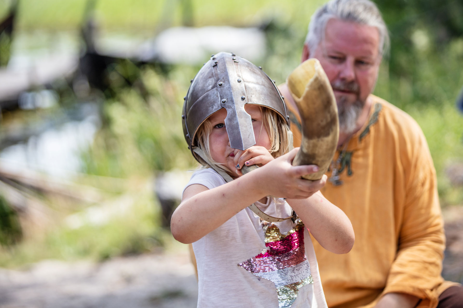 Ett barn med vikingahjälm som tutar i ett horn. En vuxen bakom barnet i medeltida kläder.
