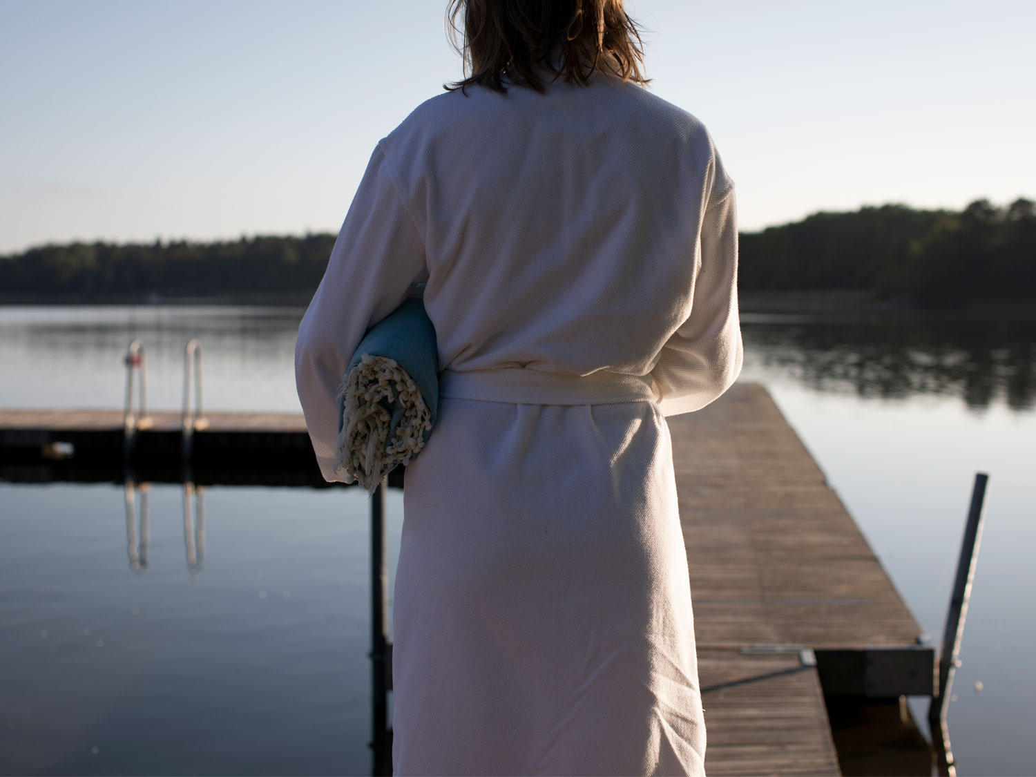 En kvinna i en vit badrock bär en turkos handduk under armen. Hon går på en brygga. Vattnet är stilla och spegelblankt.