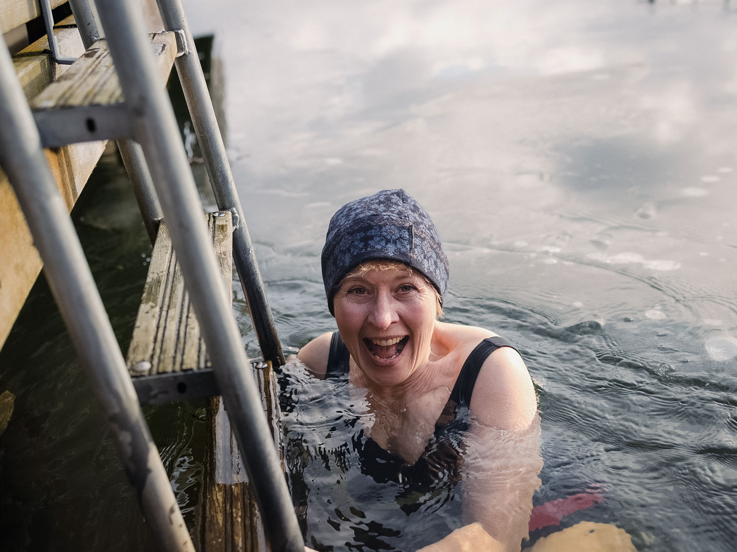En kvinna badar vinterbad. Hon håller i en stege, skrattar och har en mössa på huvudet.