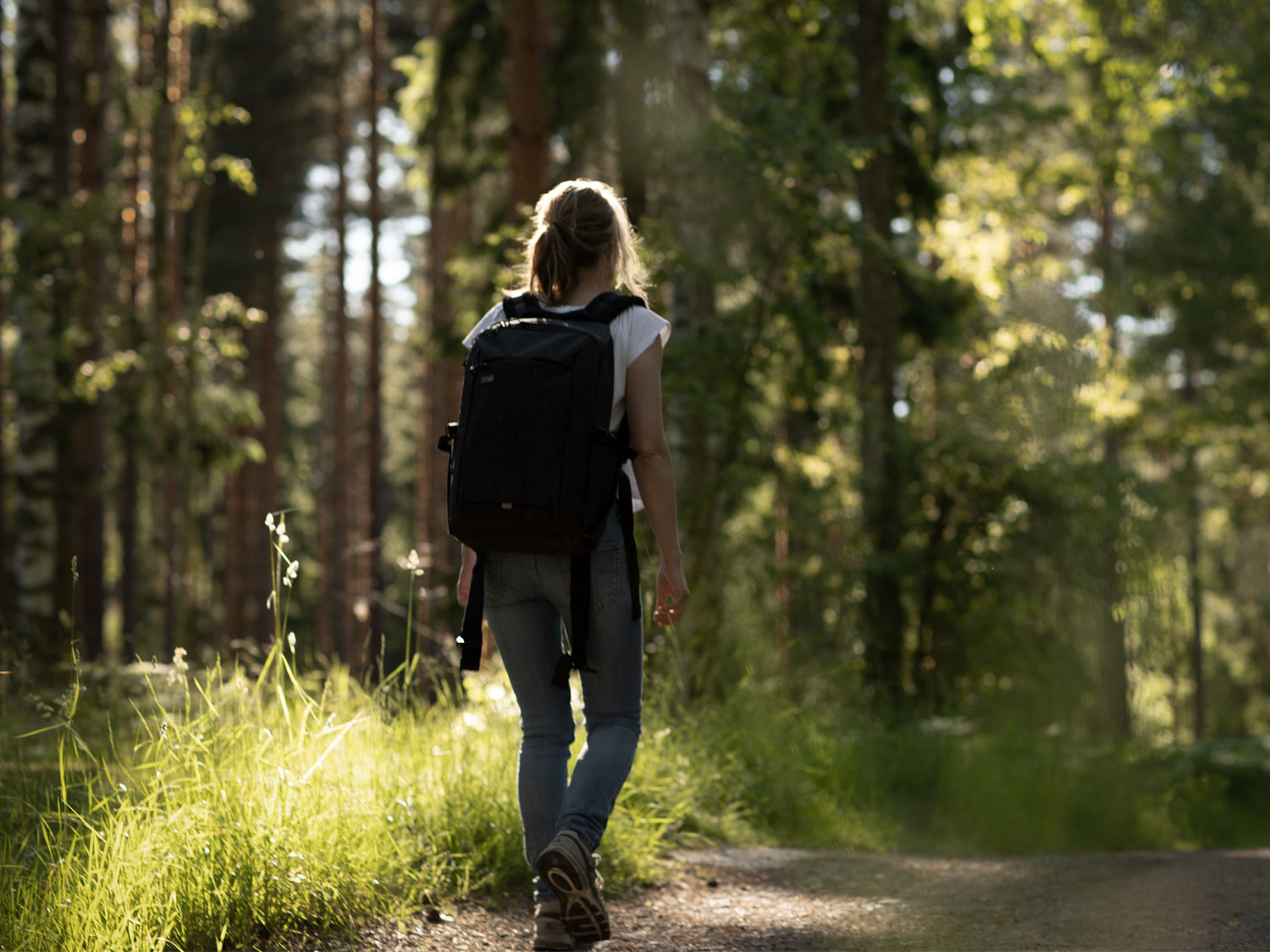 En kvinna går med i en ryggsäck på ryggen på en väg i en skog. Solen strilar genom träden.