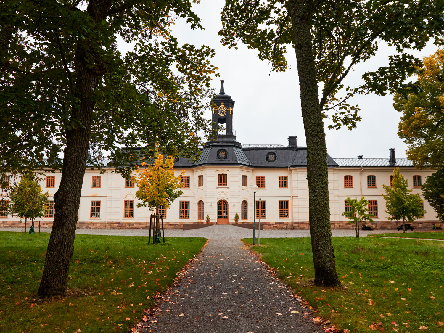 Svartsjö slott är en avlång, vitputsad byggnad med svart plåttak. I förgrunden syns en grusgång och träd.