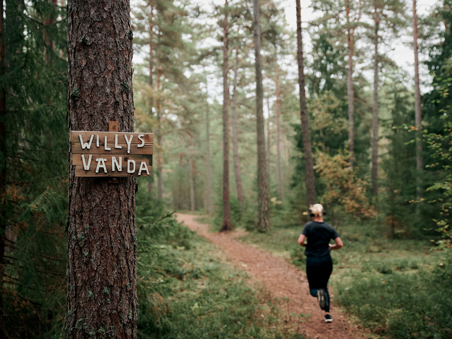 En tjej springer i träningskläder i skogen. Till vänster en trädstam med en skylt där det står Willys vända.