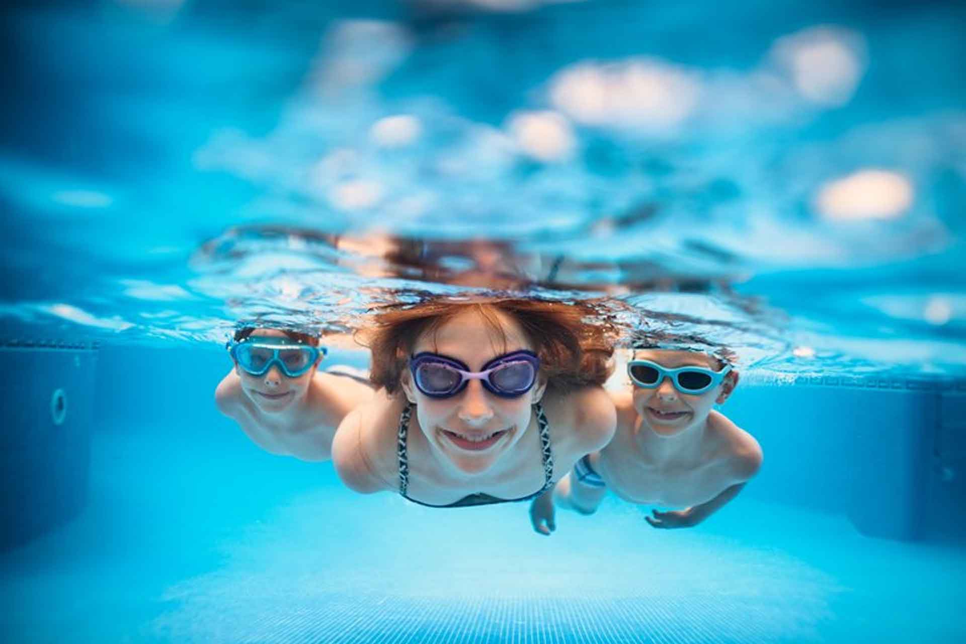 Tre barn med simglasögon som simmar i en pool under vattnet.
