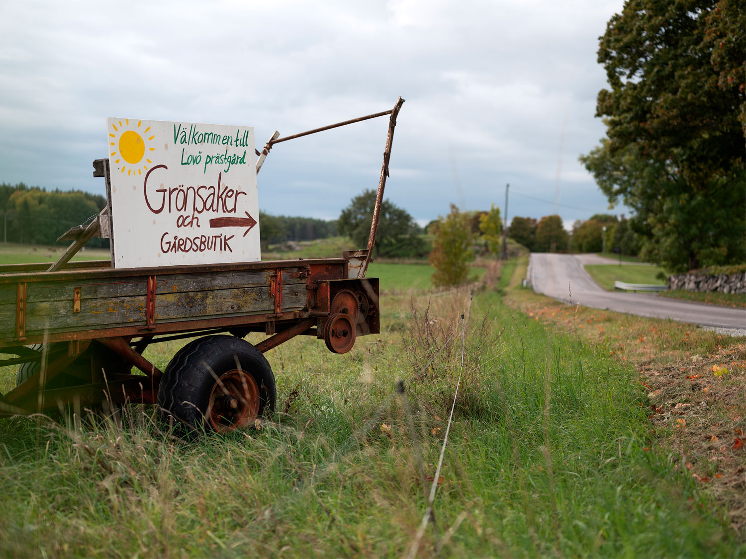 En vagn står intill en landsväg. På vagnen står en handskriven skylt med texten "Välkommen till Lovö prästgård. Grönsaker och gårdsbutik"