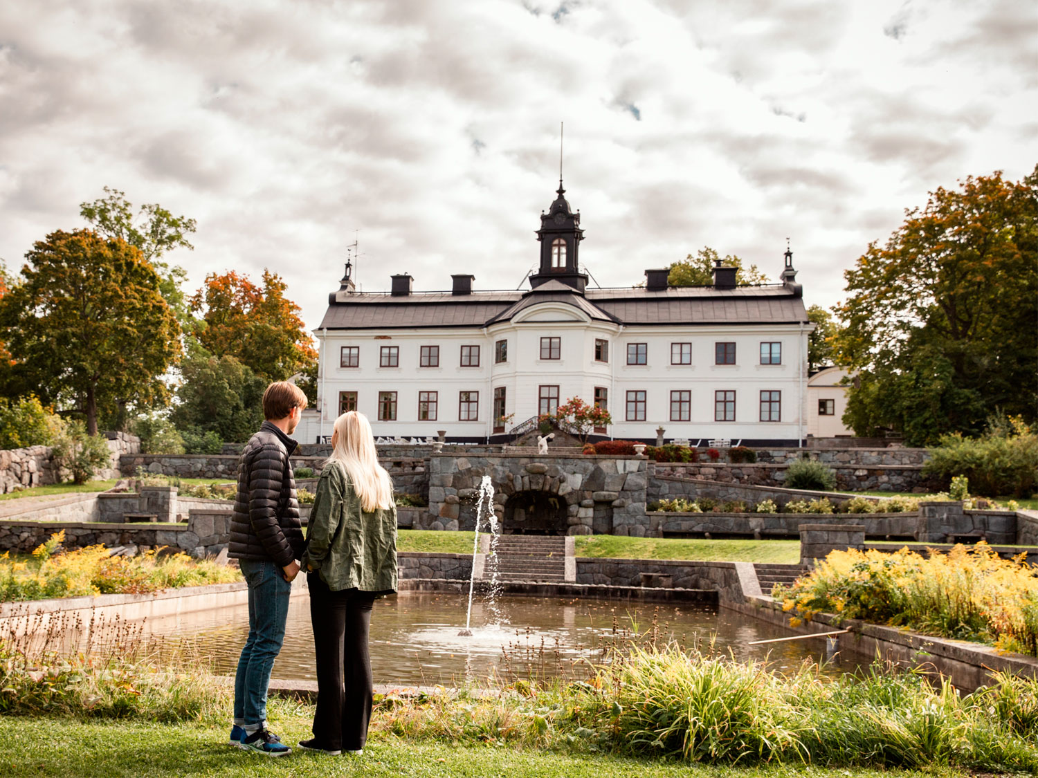 En kille och en tjej går i en slottspark. En gräsmatta syns och en fontän och statyer. I bakgrunden syns Kaggeholms slott som har en vit fasad. 