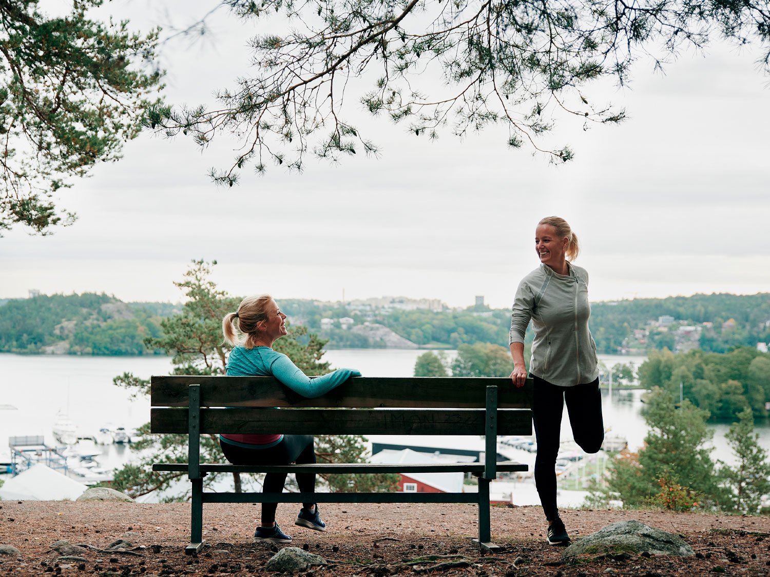 Två kvinnor i träningsläger är vid en utsiktsplats på jungfrusundsåsen där man ser jungfrusunds marina. Den ena sitter på en bänk den andra står upp och stretchar.