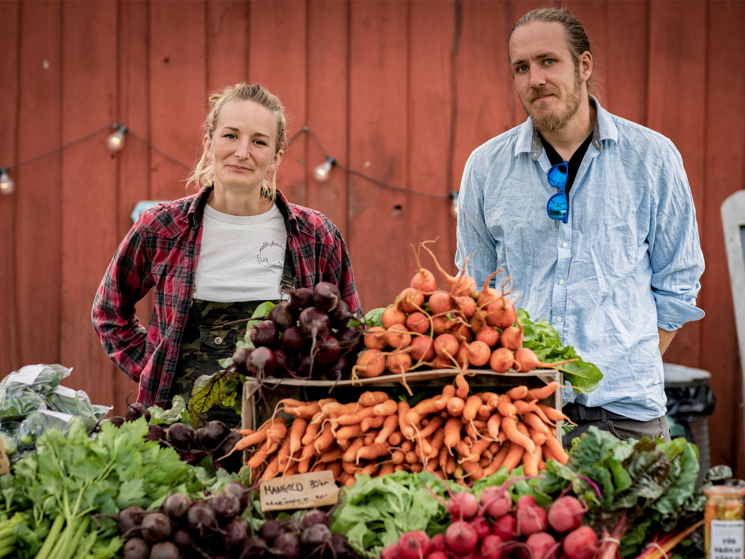 En kvinna och en man står bakom ett grönsaksstånd. Framför dem på ett bord ligger morötter, rädisor, mangold och rödbetor.