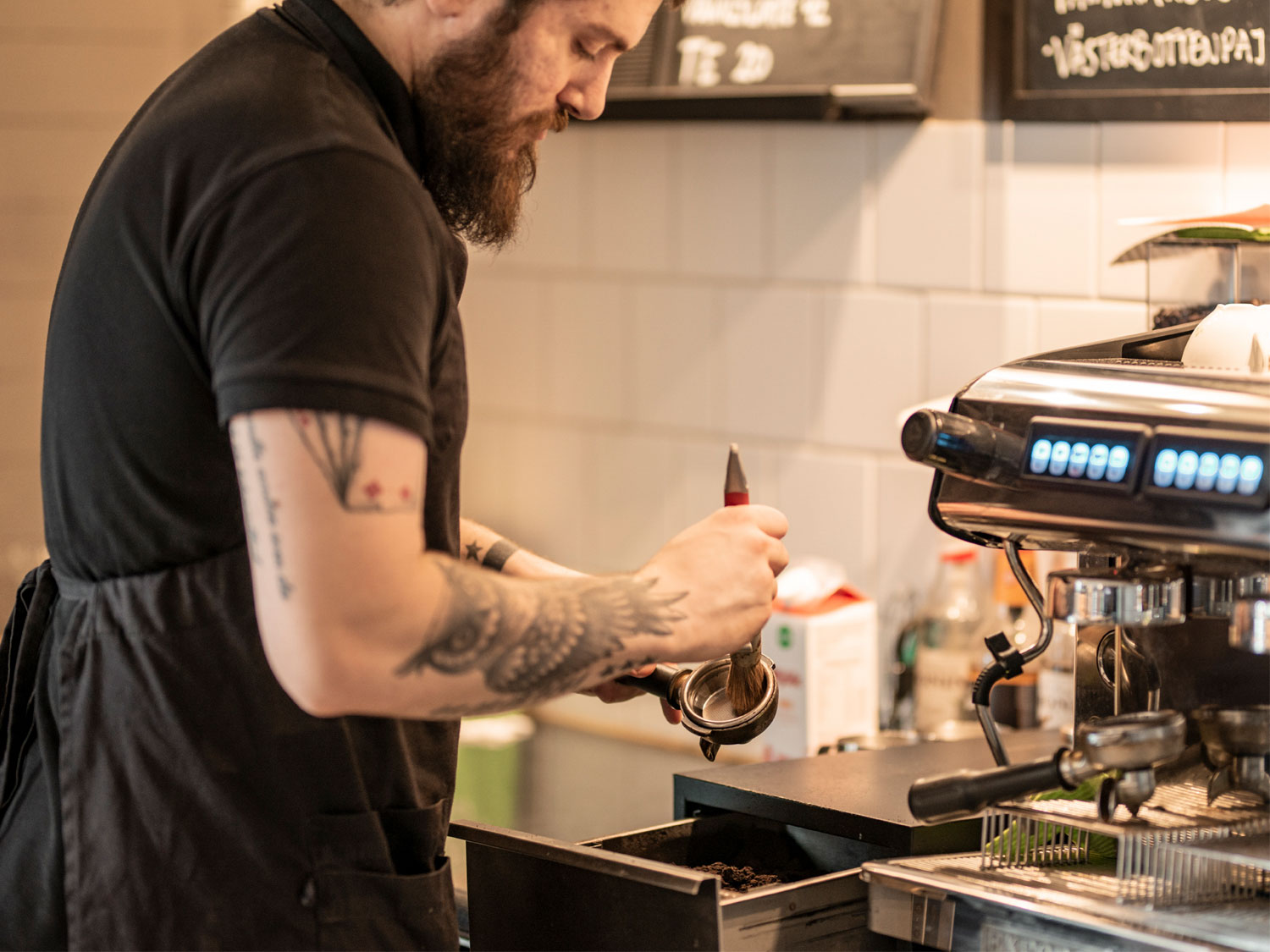 En man med tatueringar står i ett kök och gör rent en espressomaskin.