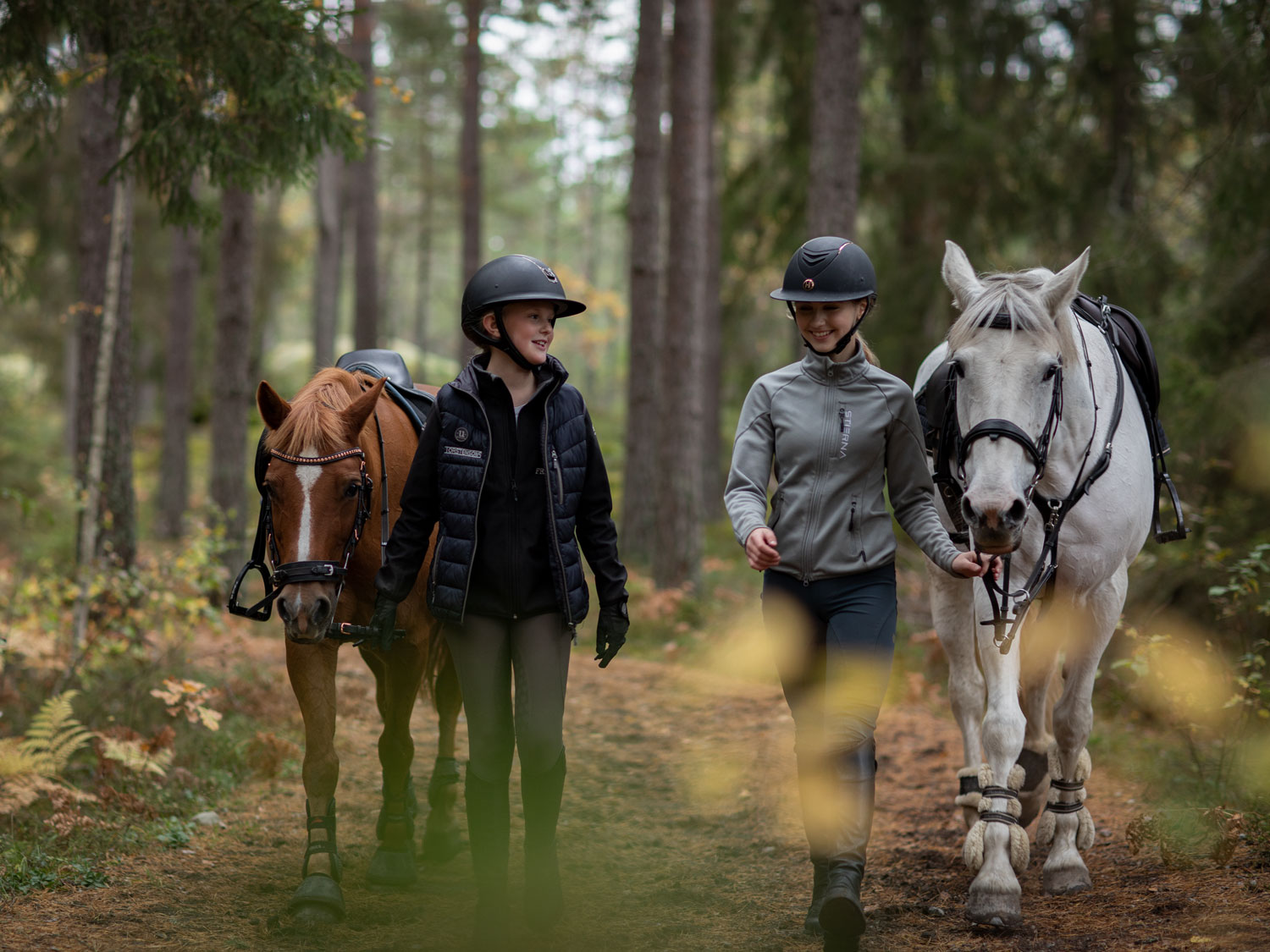 Två flickor håller i en varsin häst och går i skogen