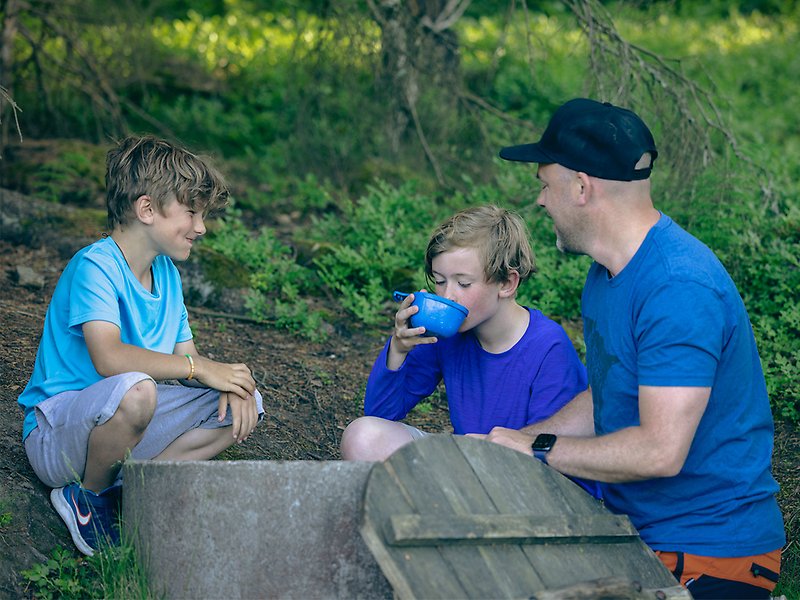 En man och två pojkar sitter runt en kallvattenkälla och dricker ur en kåsa