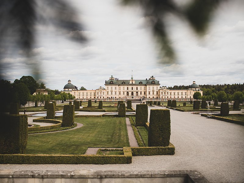Drottningholms slott syns på avstånd. Framför en del av parken med välklippta buskar och gräsmattor.