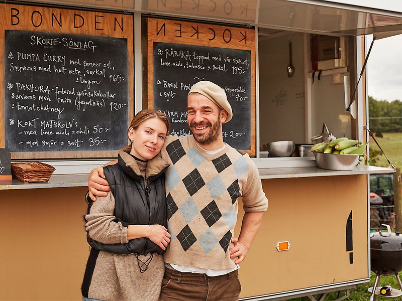 En kvinna och en man håller om varandra framför en foodtruck. Bakom den syns två griffeltavlor med menyn. 