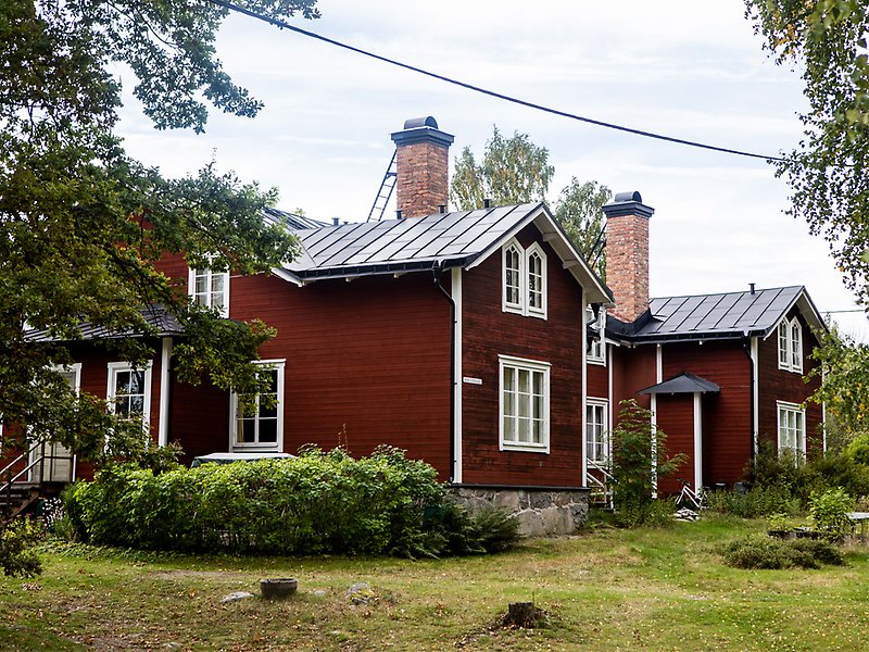 Ett stort hus i faluröd färg med vita knutar. Framför en gräsmatta och buskar.