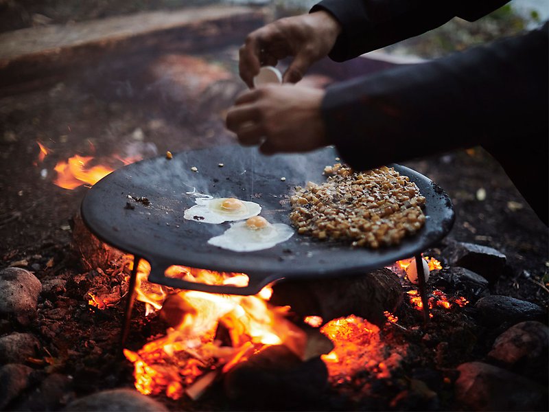 En person steker pyttipanna och ägg på en murikka över öppen eld.