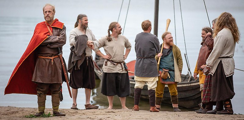 Birka - Män i vikingakläder står på en strand framför ett vikingaskepp. En man står framför dem med armarna i kors. Han har en röd mantel.