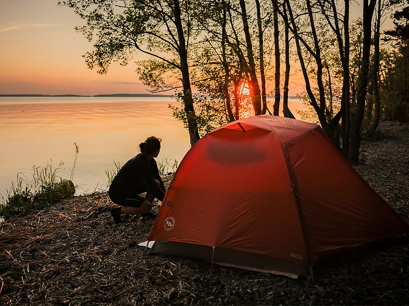 Ett rött tält står uppställt nära vattnet. Det är solnedgång. En kvinna sitter på huk bredvid tältet.