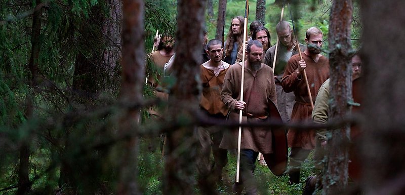 Hovgården - Män i bruna vikingakläder går i skogen. De bär på vapen, ett slags spjut.