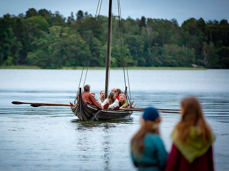 Några män ror i ett vikingaskepp. Två barn står och tittar på båten. I bakgrunden syns skog.