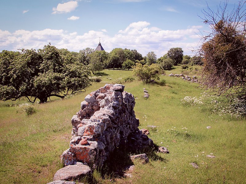 Rester av en stenmur står i en hage i Hovgården. På marken gräs i bakgrunden syns Adelsö kyrka.