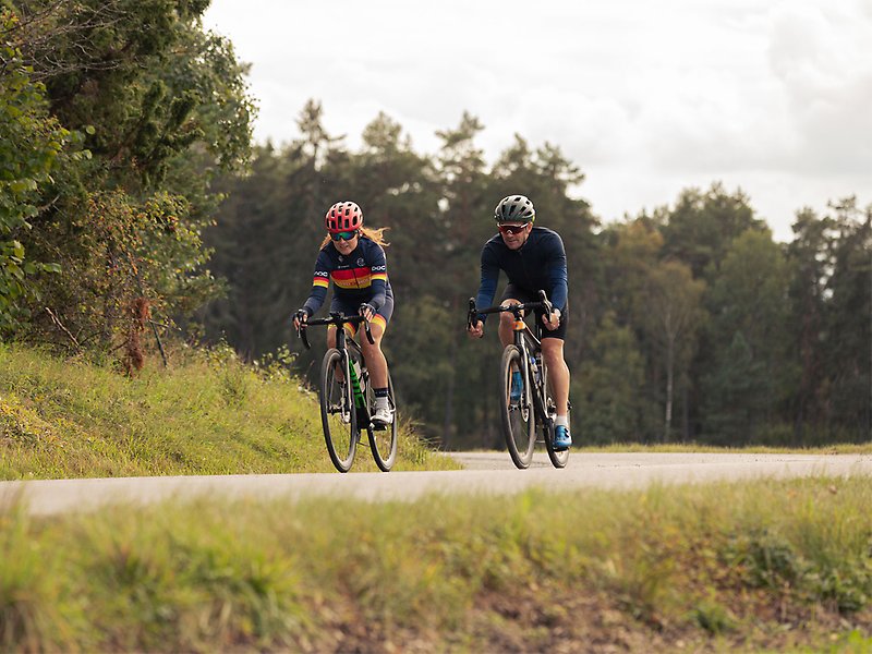 En kvinna och en man cyklar på en asfalterad väg. Runtom syns skog.