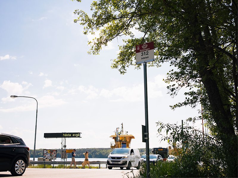 Bilar åker av en bilfärja. En busshållplatsskylt syns " Sjöängen buss 312 Adelsö" och en digital skylt som säger "nästa avgång".