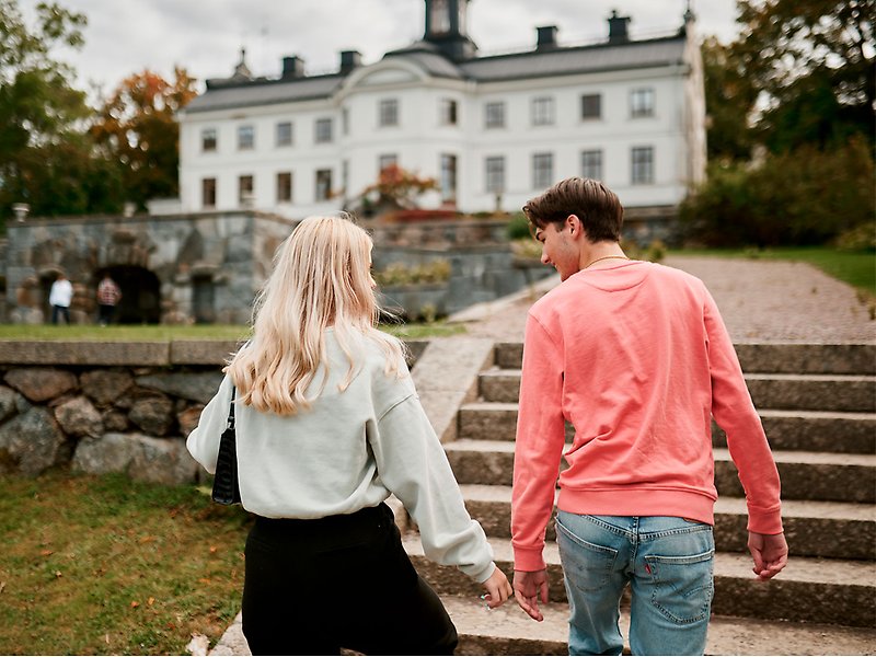 En kille och en tjej håller varandra i handen. De är på väg upp för en stentrappa. I bakgrunden syns Kaggeholms slott som har en vit fasad. 