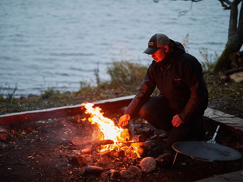 En man sitter intill en eld vid en grillplats.